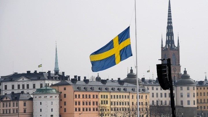 Η σημαία της Σουηδίας πίσω από κτίρια - Οι Σουηδοί θέλουν να ενταχθούν στο ΝΑΤΟ αλλά όχι τόσο βιαστικά