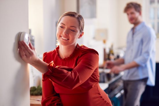 Pregnant Couple With Woman With Prosthetic Arm Turning Down Heating Control In Kitchen Together